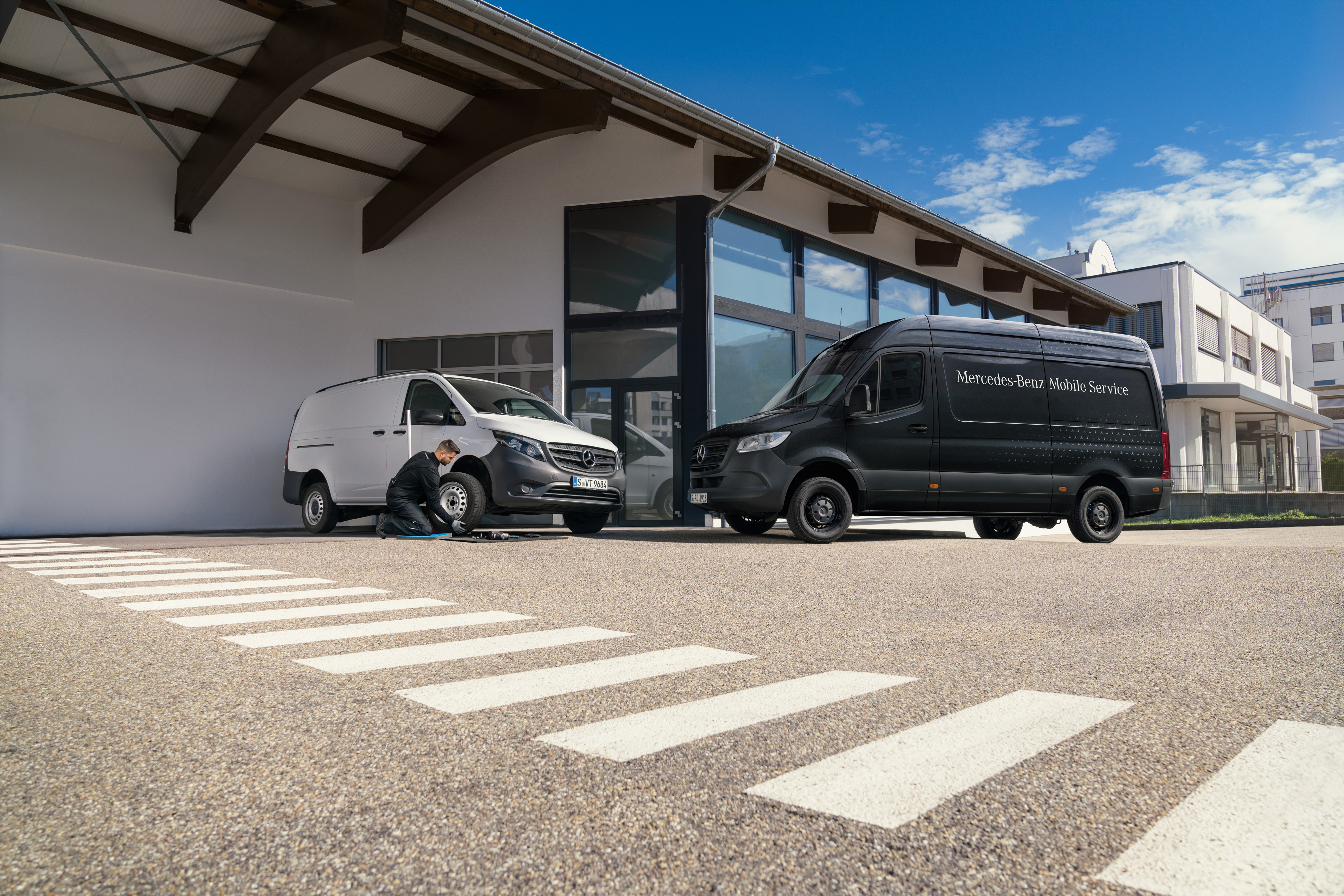 Mercedes-Benz | Mobile Service | Bremsanlage Ein Techniker wechselt einen Reifen an einem weißen Mercedes-Benz Vito vor einem Ausstellungsraum.