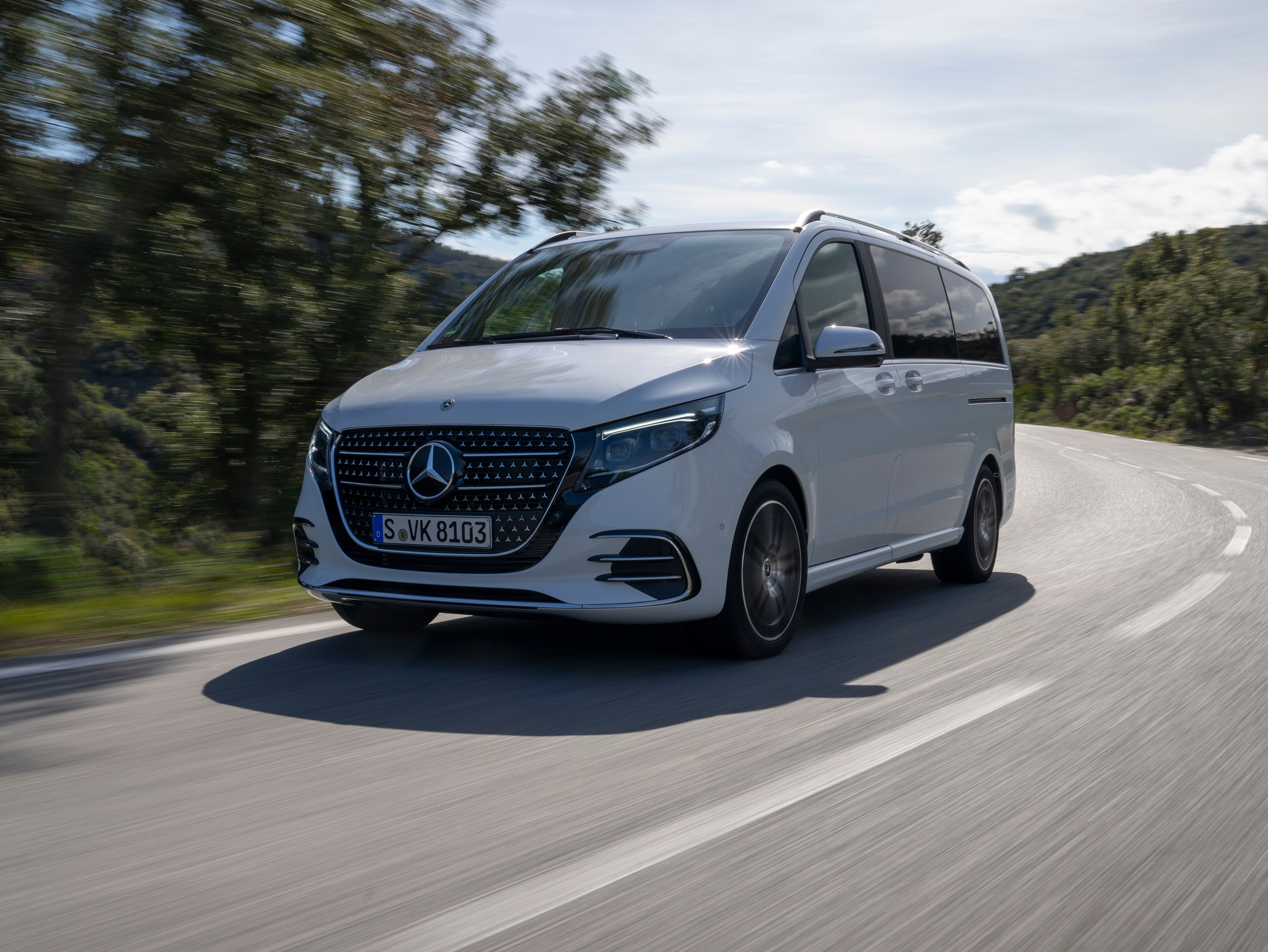 Eine weiße V-Klasse von Mercedes-Benz fährt auf einer sonnigen Landstraße an Bäumen entlang.
