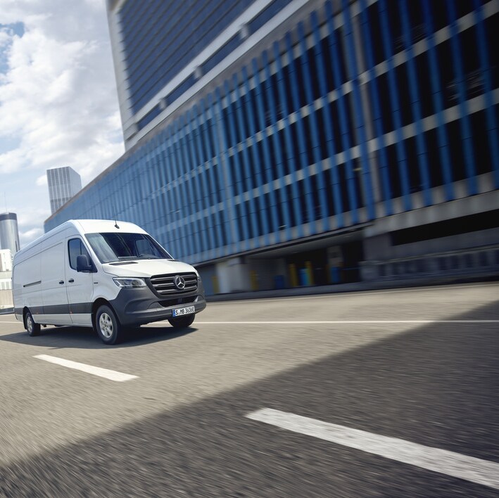 Ein weißer eSprinter Kastenwagen fährt auf einer Straße in einer urbanen Umgebung.
