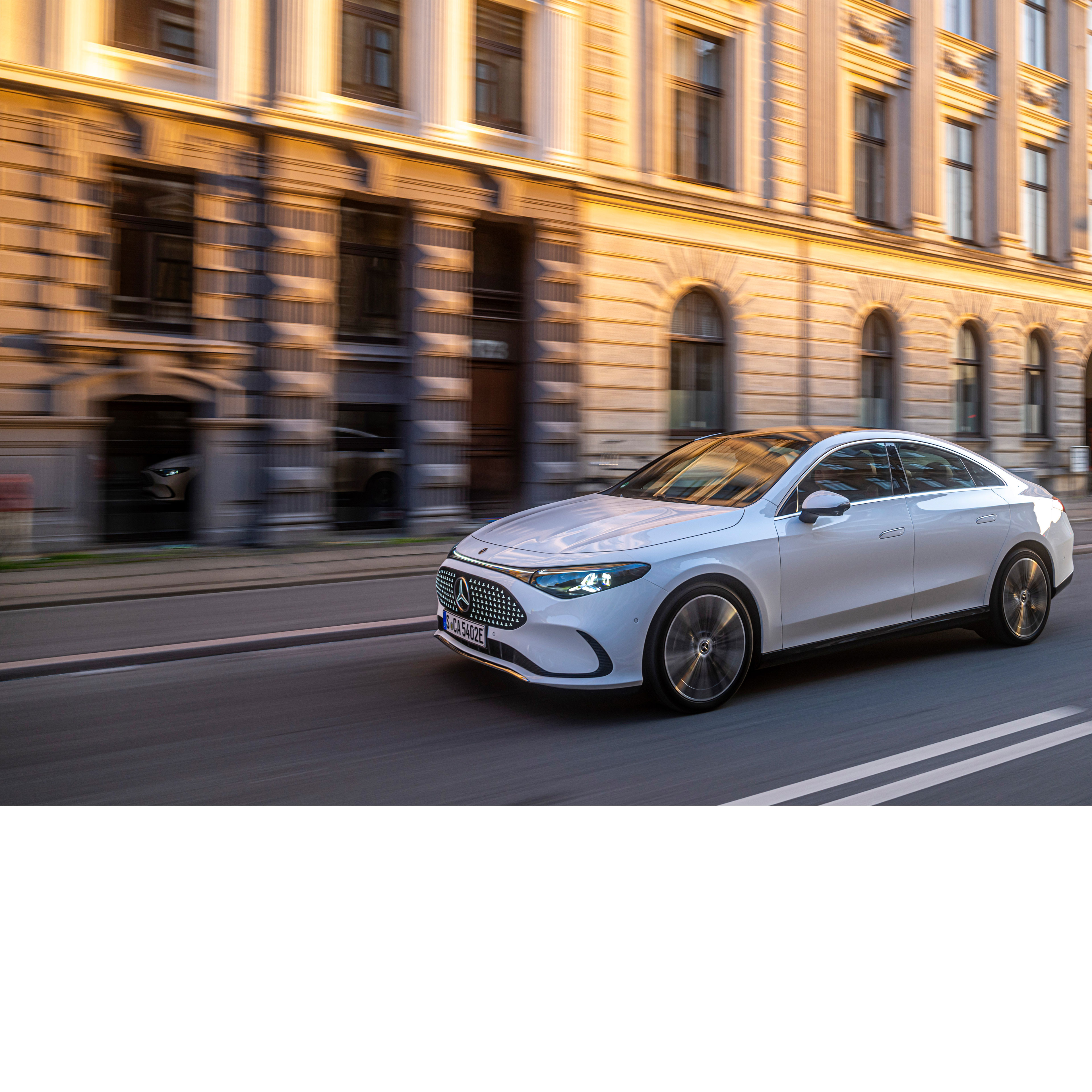 Automatisiertes Fahren & Assistenzsysteme | Mercedes-Benz Ein heller Mercedes-Benz fährt an einem steinernen Gebäude bei untergehender Sonne entlang.