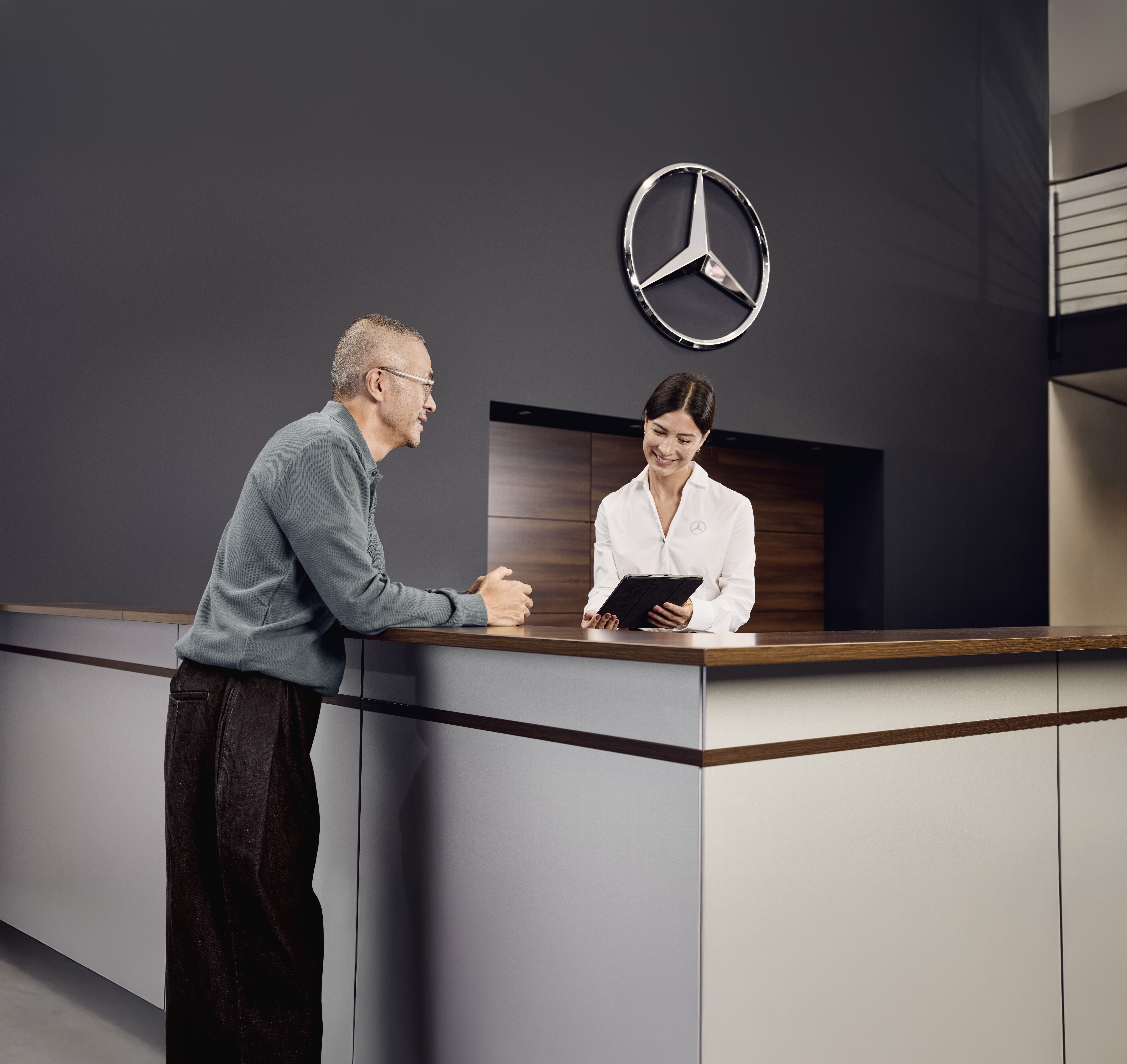 Ersatzteile | Mercedes-Benz Empfangsbereich der Serviceabteilung eines Mercedes-Benz-Händlers, wo ein Kunde mit einem Serviceberater am Empfang die Einzelheiten der Fahrzeugwartung bespricht.